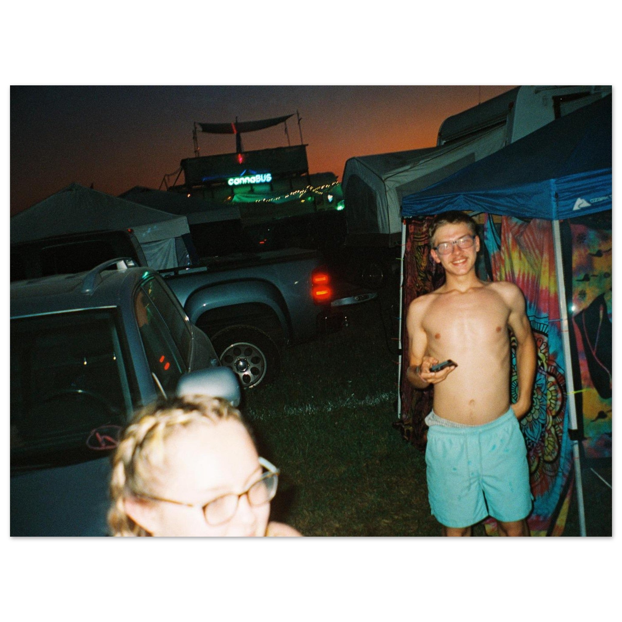 Two people at a nighttime outdoor event with tents and vehicles.
