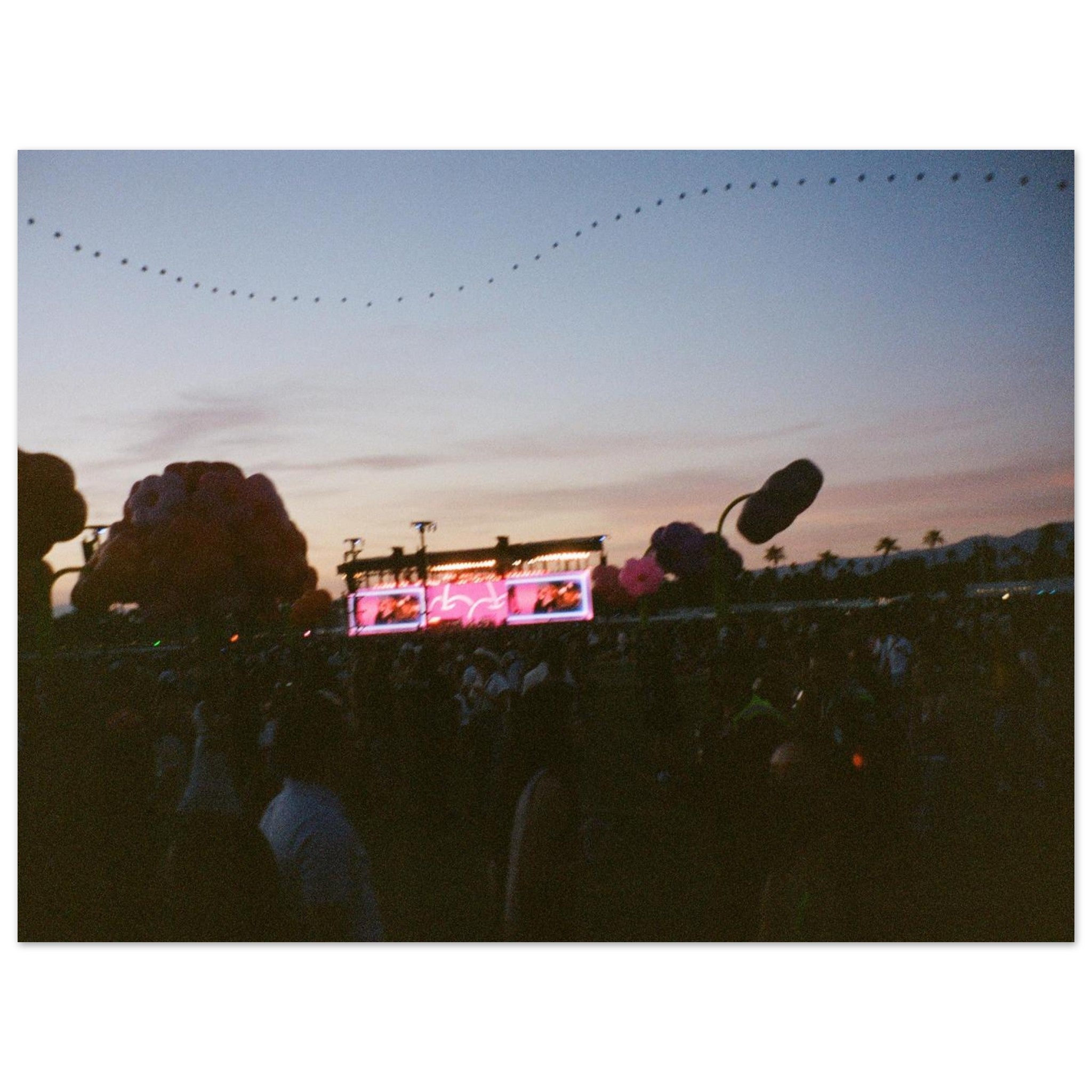 People at a concert with a stage and screens in the background during sunset.